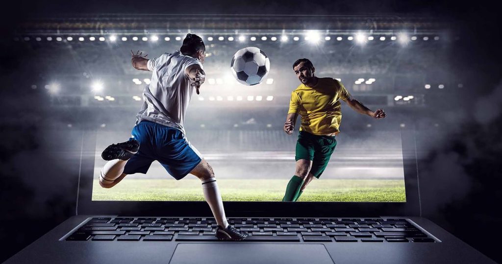 An advertisement for a sports betting service. The image shows two soccer players, one in a white jersey and blue shorts, and the other in a yellow jersey and green shorts, mid-game. They appear to be leaping out of a laptop screen, which is in the foreground. The background is a brightly lit stadium at night.