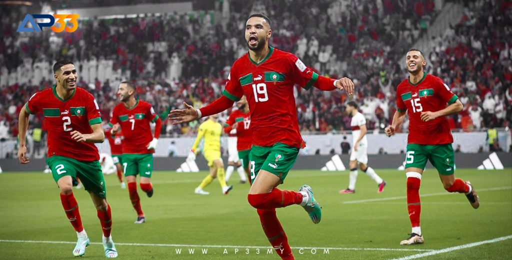 An advertisement for the online sportsbook AP33, featuring a soccer player from the Morocco national team in a red jersey with the number 19, celebrating a goal. He is yelling with his arms outstretched, with other teammates running to join him. The background is a blurry stadium with a crowd of spectators.
